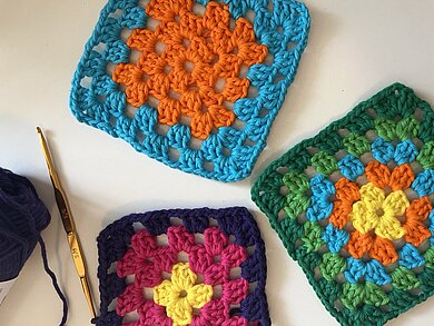 Mehrere bunte Granny Squares mit einer Häkelnadel liegen auf dem Tisch. 