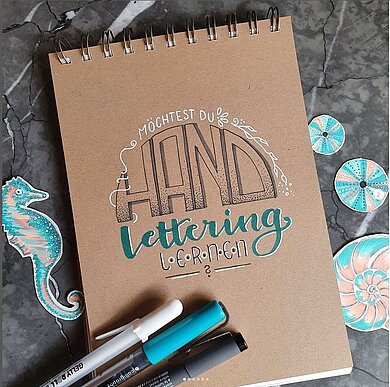 Blick auf einen Block mit verschiedenen Handlettering-Schriften, 3 verschiedenfarbige Stifte, ein gemaltes Seepferdchen und Muscheln