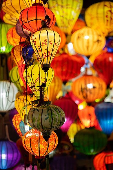 Das Bild ist im Hochformat. Es ist gefüllt mit runden und teils länglichen vietnamesische Lampen. Die Lampen sind überwiegend gelb und orange, mit wenigen in grün, blau und weiß. Die Lampions haben zarte Blumenmuster oder sind einfarbig. 
