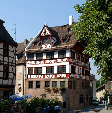 Fachwerkhaus mit steilem Dach und mehreren Fensterreihen an einer Straßenecke, umgeben von Bäumen und weiteren Gebäuden
