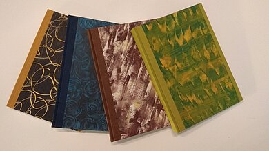 Abgebildet sind vier gebundene Bücher mit unterschiedlichen grafischen Mustern und Farben.