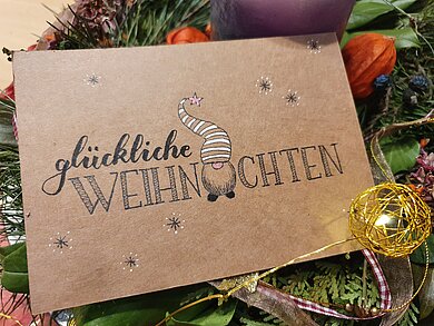 braune Weihnachtskarte mit Handlettering-Buchstaben gemalt und im Zentrum ein Weihnachtsmann, im Hintergrund ist ein Weihnachtsgesteck zu erkennen. 