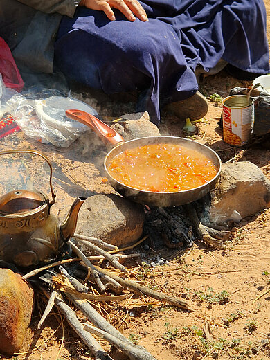 Pfanne mit kochender Suppe auf offenem Feuer aus Ästen, daneben Teekessel und Dose, Person mit blauem Gewand sitzt im Hintergrund.