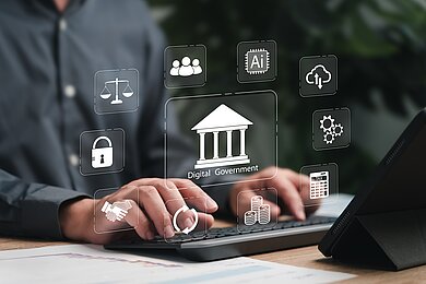 Eine Person, die an einem Schreibtisch auf einer Tastatur arbeitet. Über der Szene sind verschiedene transparente, digitale Symbole eingeblendet. Im Mittelpunkt befindet sich ein Symbol eines Regierungsgebäudes mit der Aufschrift „Digital Government“. Rundherum sind weitere Icons angeordnet, die typische Bereiche der digitalen Verwaltung darstellen: Waage, Personengruppe, KI-Chip, Cloud mit Pfeilen, Zahnräder, ein Steuerrechner, Münzen, Kreispfeile, Handschlag, Schloss.
