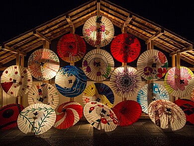 Das Bild zeigt bunte japanische Papierschirme mit der Oberseite zum Bild-Betrachter gerichtet. Die aufgespannten Schirme sammeln sich schwebend unter einem Holzbaldachin.