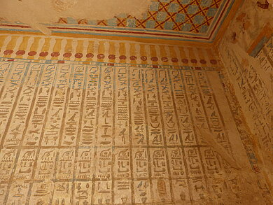 Wand mit senkrechten Reihen altägyptischer Hieroglyphen und farbigen Mustern am oberen Rand